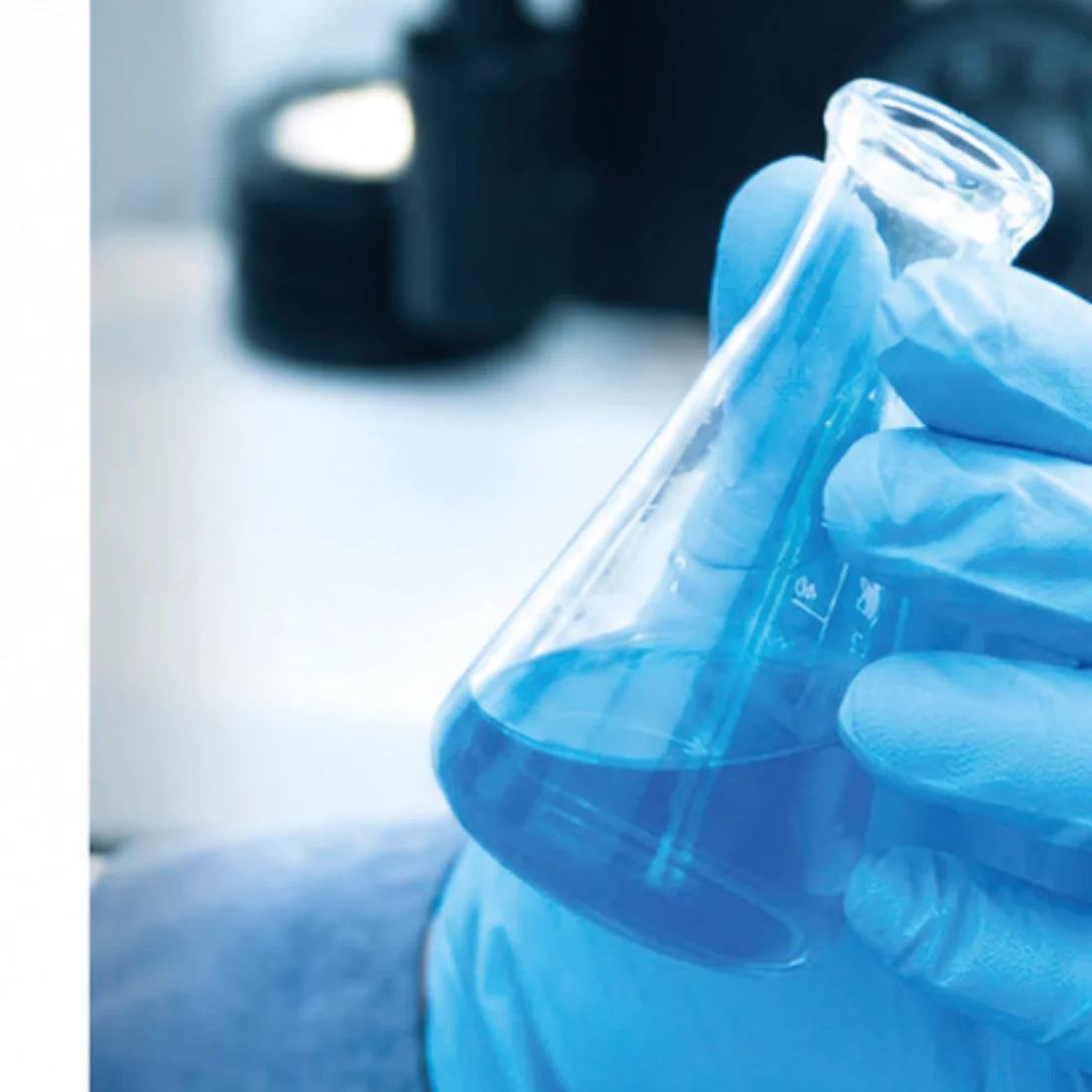 Mano con guanto in nitrile blu che regge una beuta con liquido azzurro, su un bancone di laboratorio con capsule Petri.